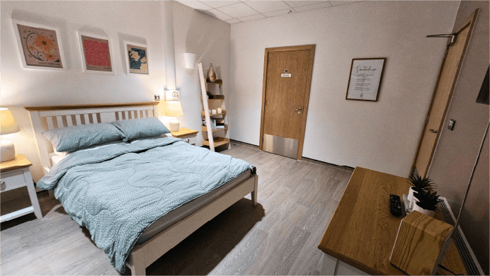 A Double Bed with patterned duvet, flanked by bedside tables furnished with soft glowing lamps. Three flower illustrations hang above with a bookcase to one side offering lit candles and books. Opposite, a chest of drawers with a digital radio.