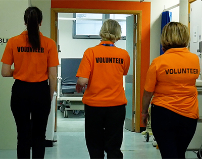 The backs of three women wearing orange tops with volunteer written on them, walking down a clinical corridor