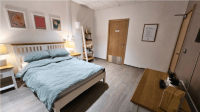 A Double Bed with patterned duvet, flanked by bedside tables furnished with soft glowing lamps. Three flower illustrations hang above with a bookcase to one side offering lit candles and books. Opposite, a chest of drawers with a digital radio.